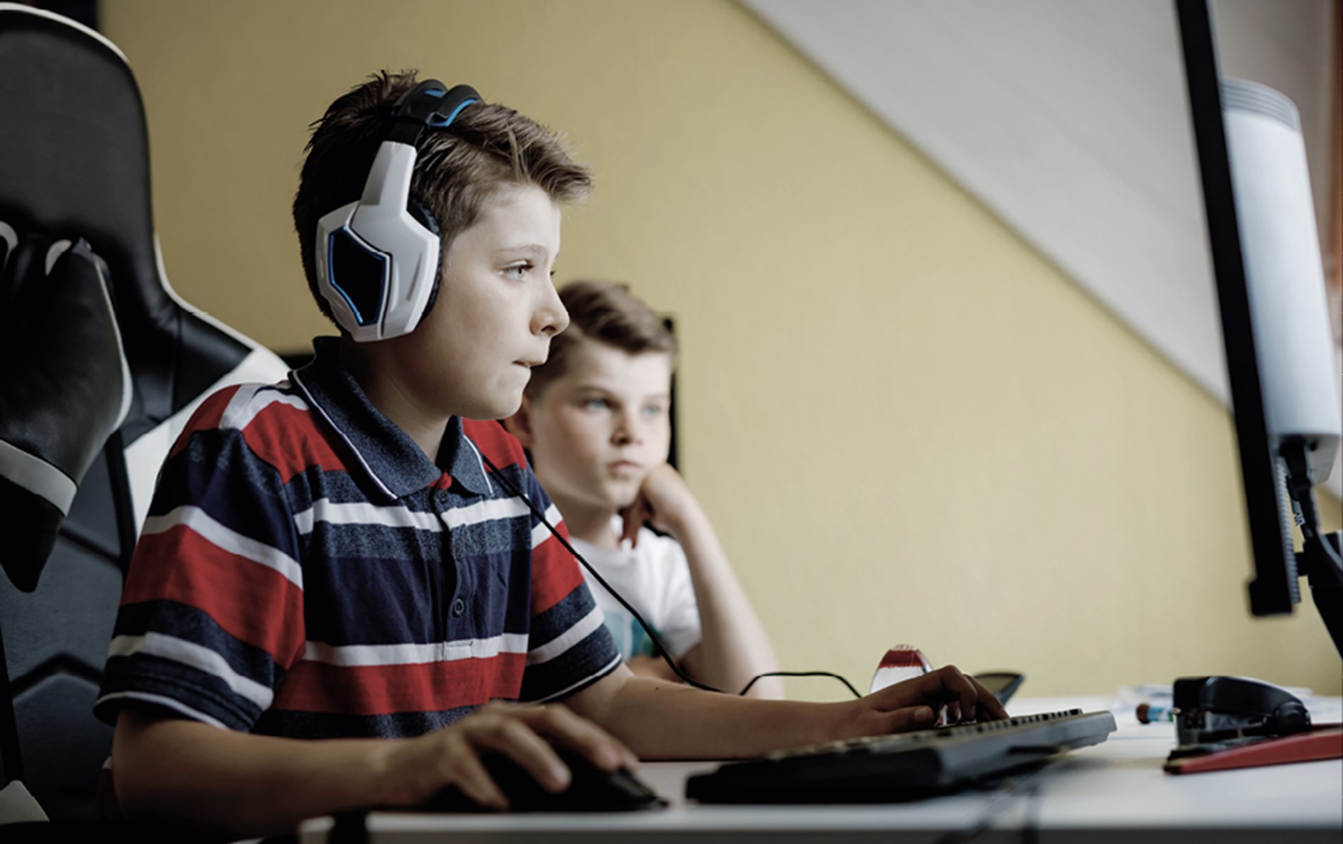 Ein Kind sitzt vor einem Computer und trägt dabei ein weißes Headset. Ein Junge sitzt daneben und schaut zu.