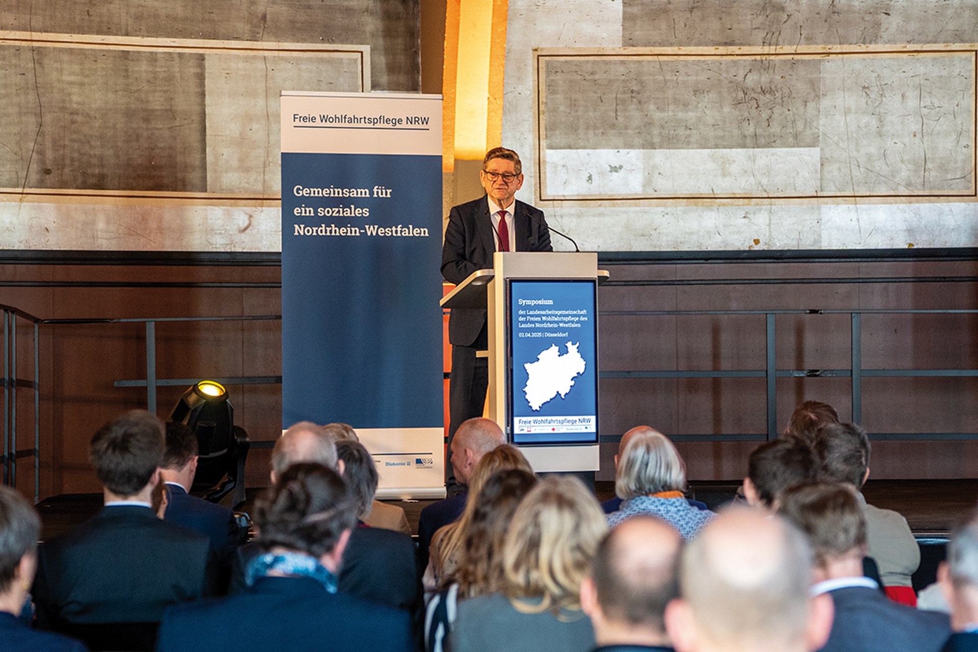 Ein NRW-Landtagsabgeordneter, der auf einer Bühne vor einem Rednerpult steht und beim Symposium der LAG Freie Wohlfahrtspflege NRW vor dem Publikum ein Grußwort hält