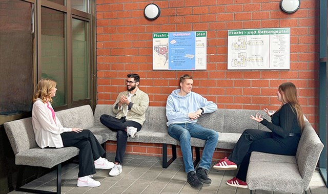 Je zwei junge Frauen und Männer sitzen zusammen auf der grauen 'Plauderbank' und sprechen miteinander.