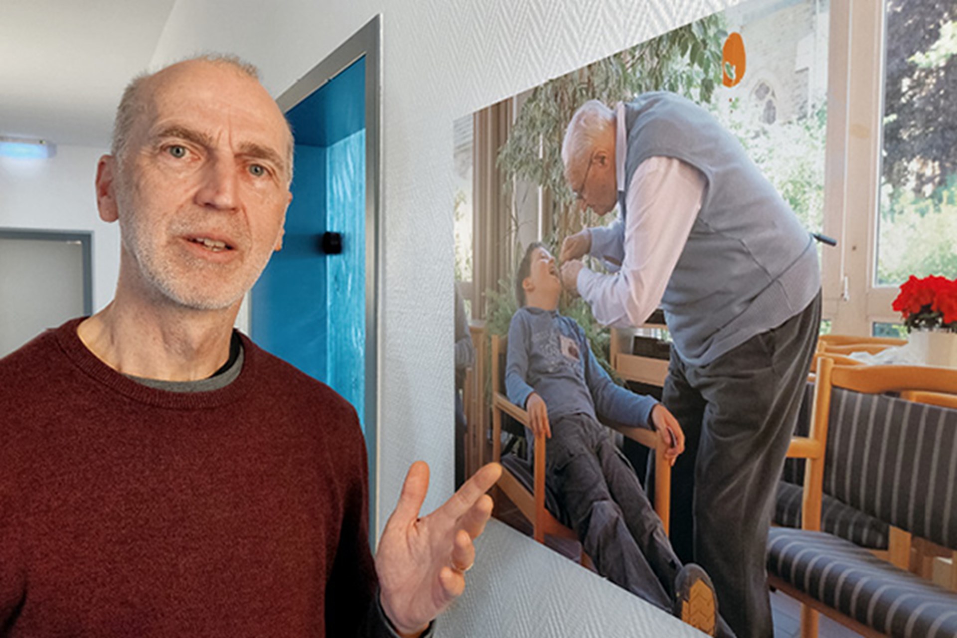 Ein Mann in einem roten Pullover, der auf einem Flur steht und auf ein an der Wand hängendes Foto deutet. Auf dem Foto sind ein auf einem Stuhl sitzender Junge und ein Senior zu sehen, der sich über den Jungen beugt.
