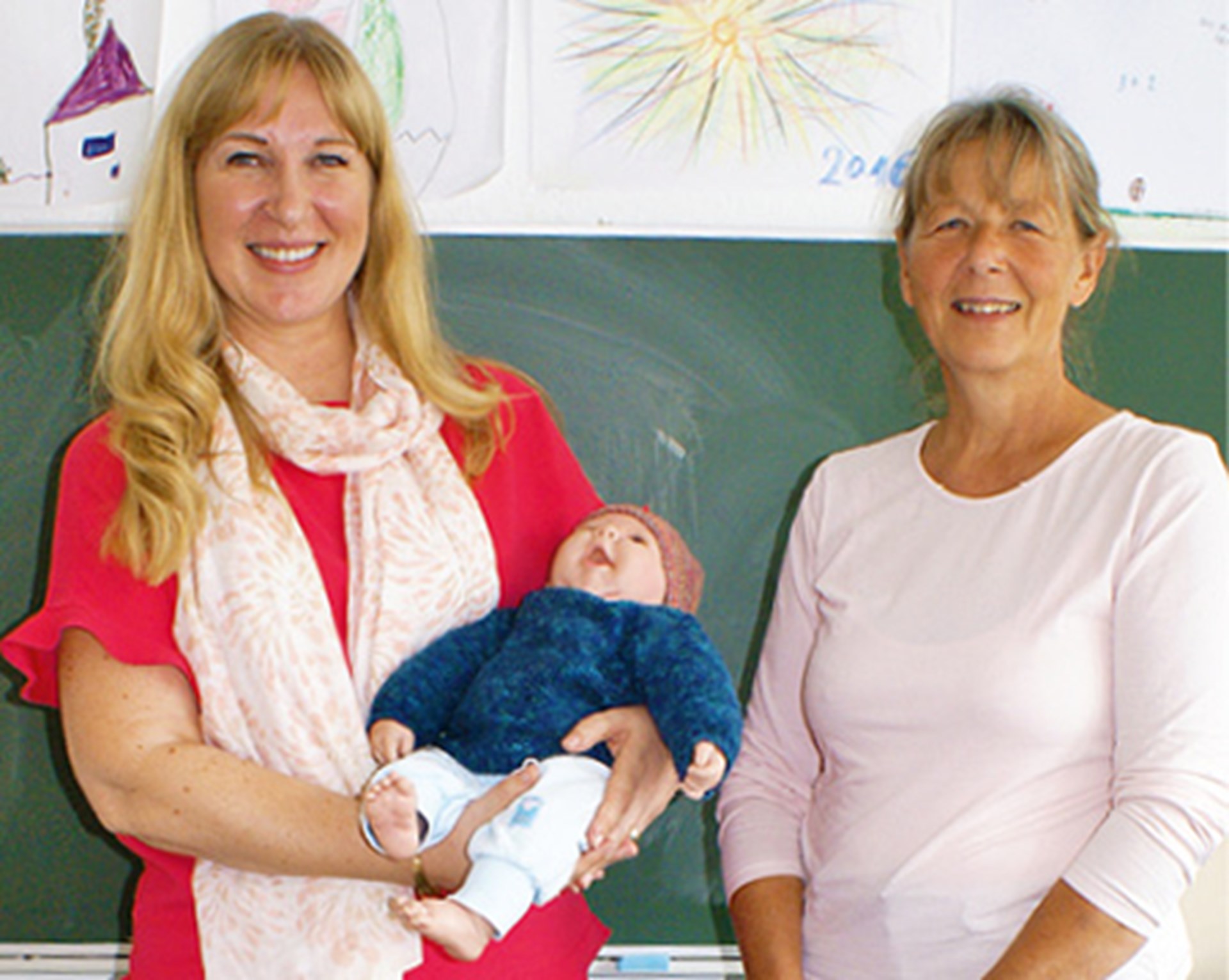 Zwei Frauen stehen vor einer grünen Tafel. Eine Frau trägt eine Babypuppe im Arm.