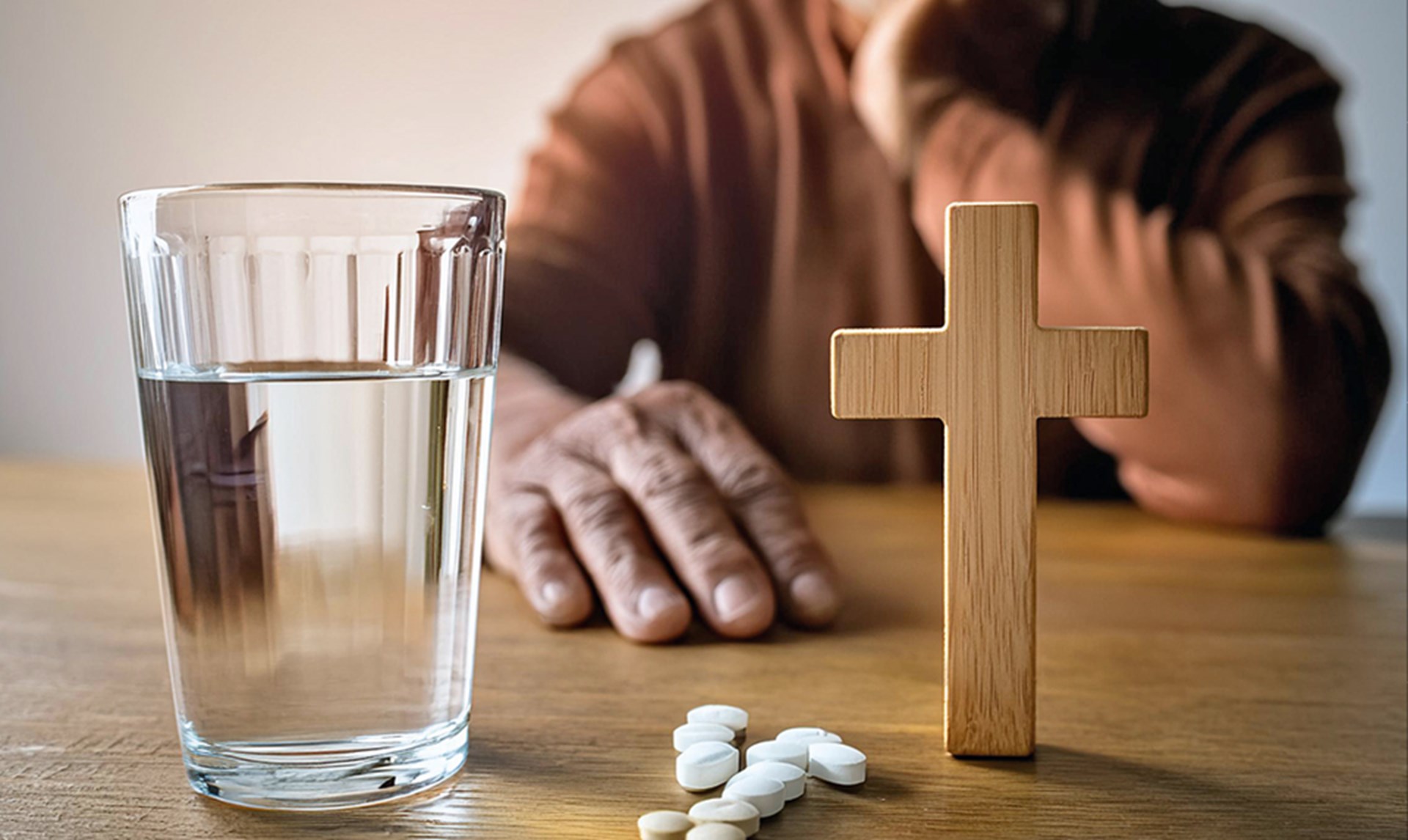 Ein Senior sitzt an einem Holztisch. Vor ihm sieht man ein Glas mit Wasser, einige Tabletten und ein kleines Holzkreuz.