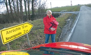 Eine Frau in einer roten Jacke steht vor ihrem roten Dienstwagen an einer Landstraße. Neben ihr ist ein Schild mit zwei Orts- und Kilometerangaben zu sehen, im Hintergrund erstreckt sich eine Wiesenlandschaft.