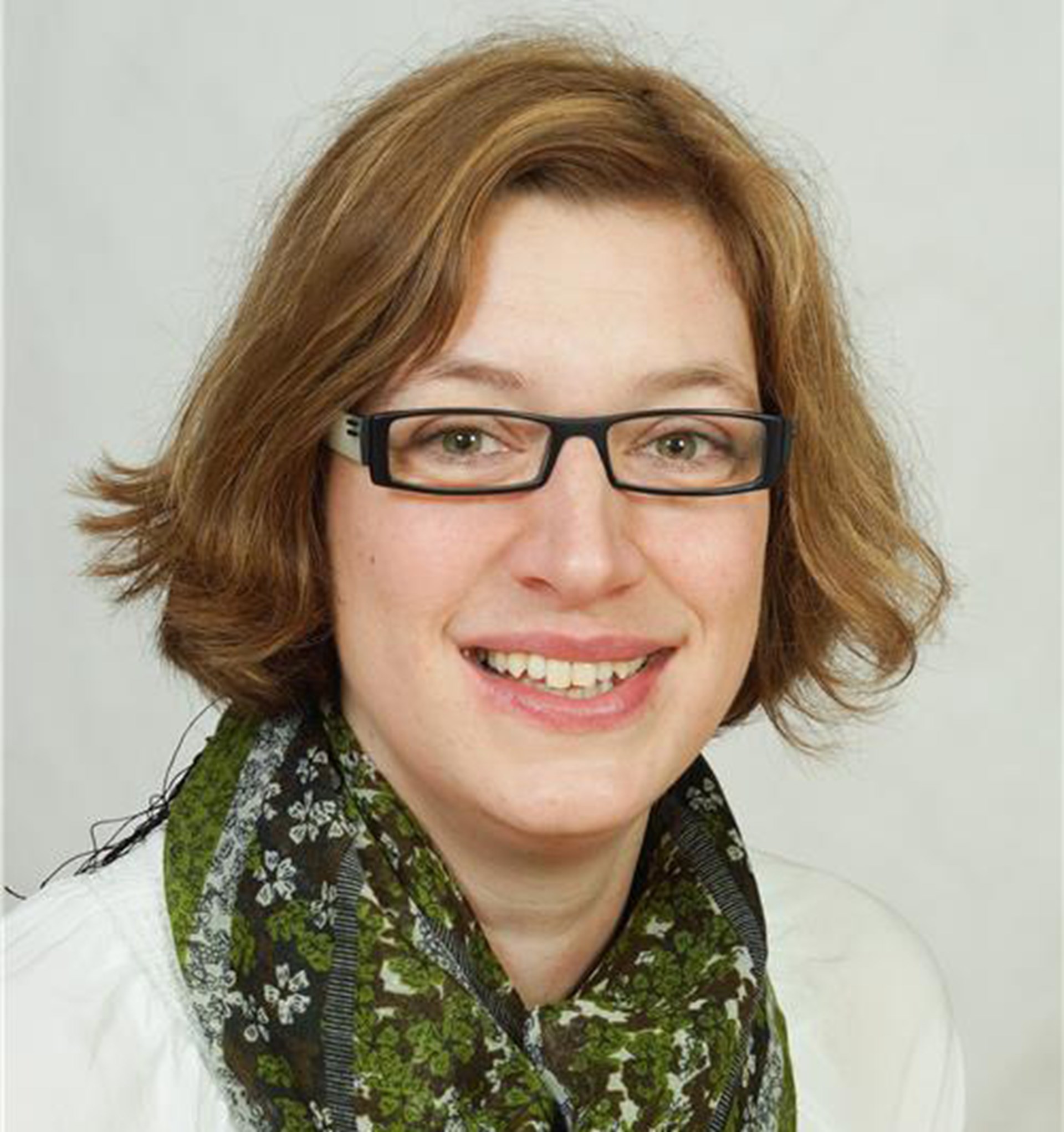 Lächelnde Frau mit blonden Haaren und Brille, die einen grünen Schal und ein weißes T-Shirt trägt