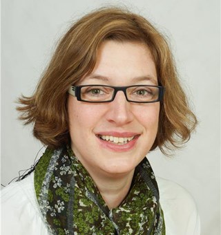 Lächelnde Frau mit blonden Haaren und Brille, die einen grünen Schal und ein weißes T-Shirt trägt