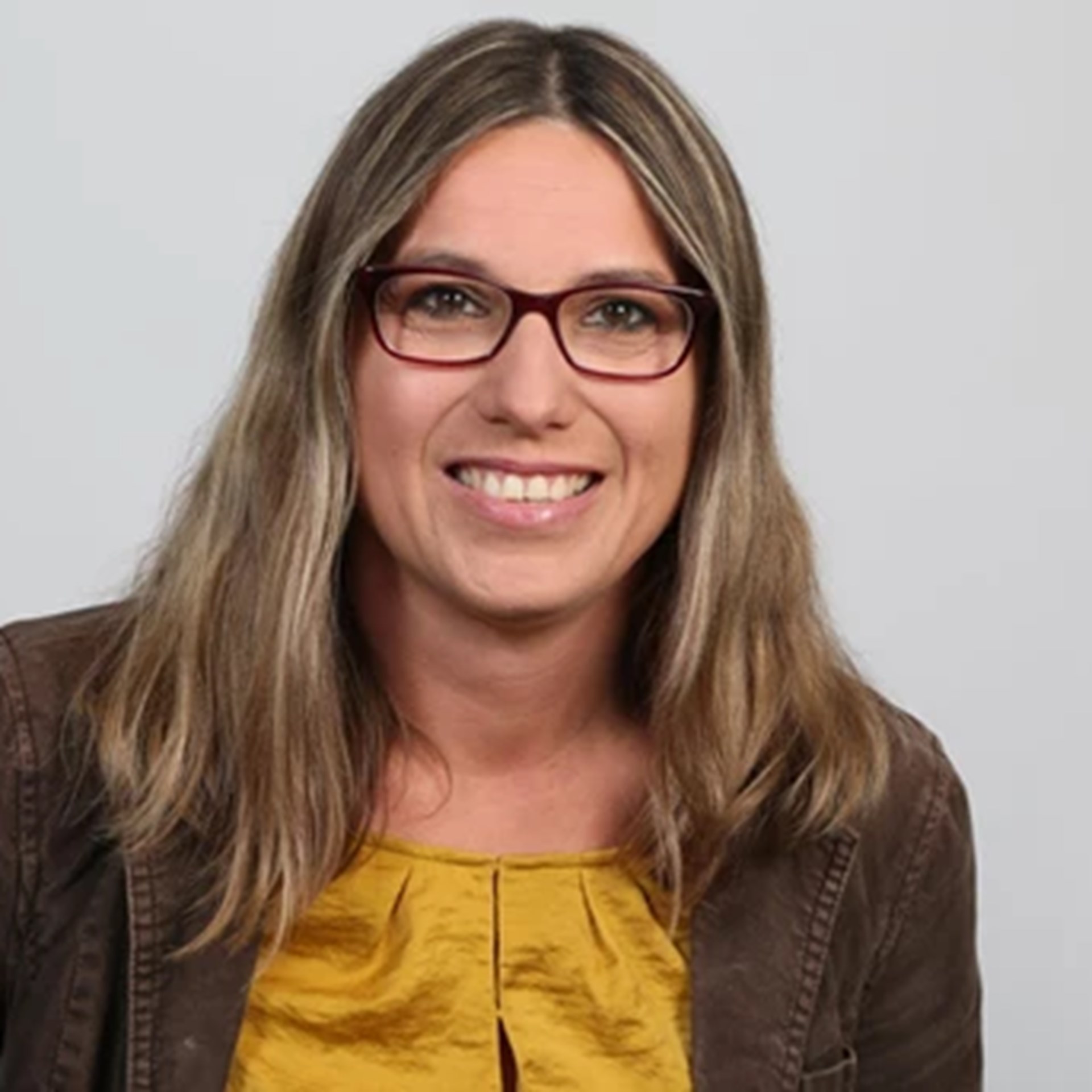 Lächelnde Frau mit schulterlangen, blonden Haaren und Brille, die eine braune Jeansjacke und ein gelbes Shirt trägt