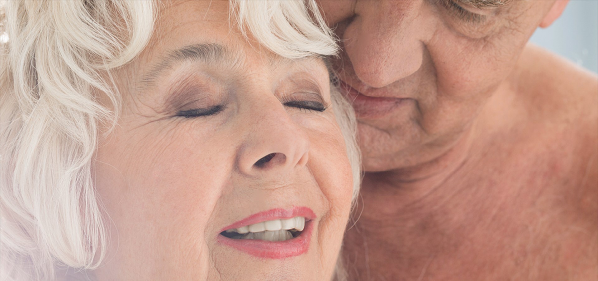 Eine intime Szene, in der sich ein Senior mit nacktem Oberkörper einer Seniorin zärtlich von hinten annähert