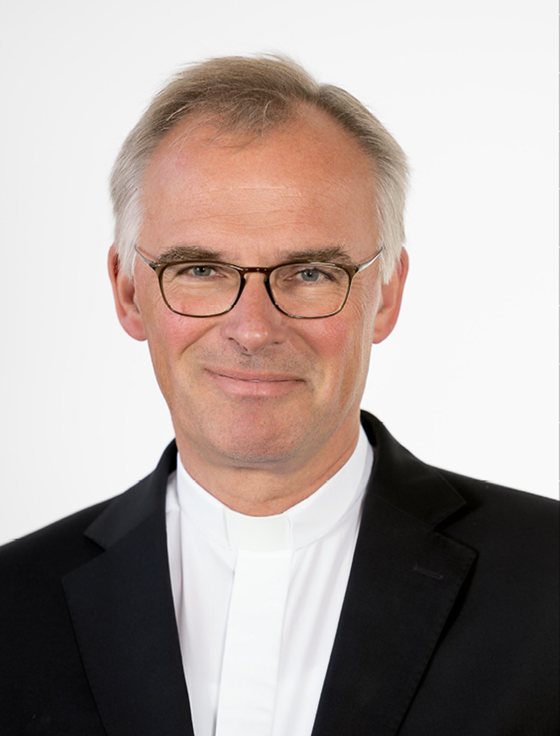 Lächelnder Mann mit grauen Haaren und Brille, der ein weißes Hemd mit Piuskragen (Kollar) und eine dunkelblaue Anzugjacke trägt
