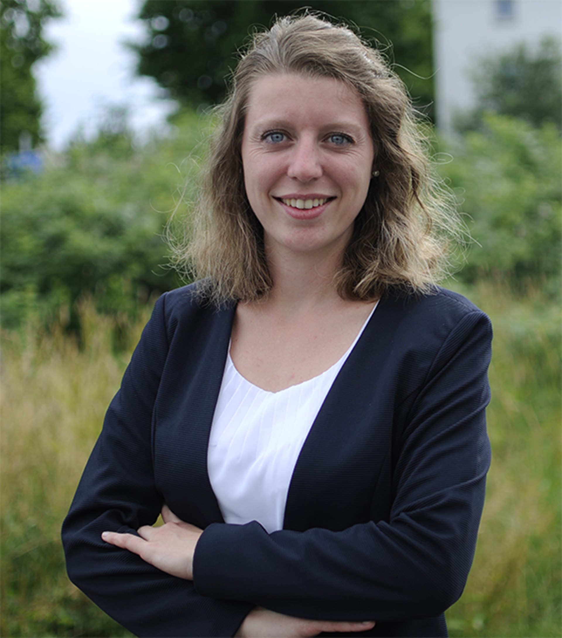 Lächelnde, junge Frau mit langen, blonden Haaren, die ein weißes Shirt mit einer blauen Jacke trägt und mit verschränkten Armen in einer Grünanlage steht