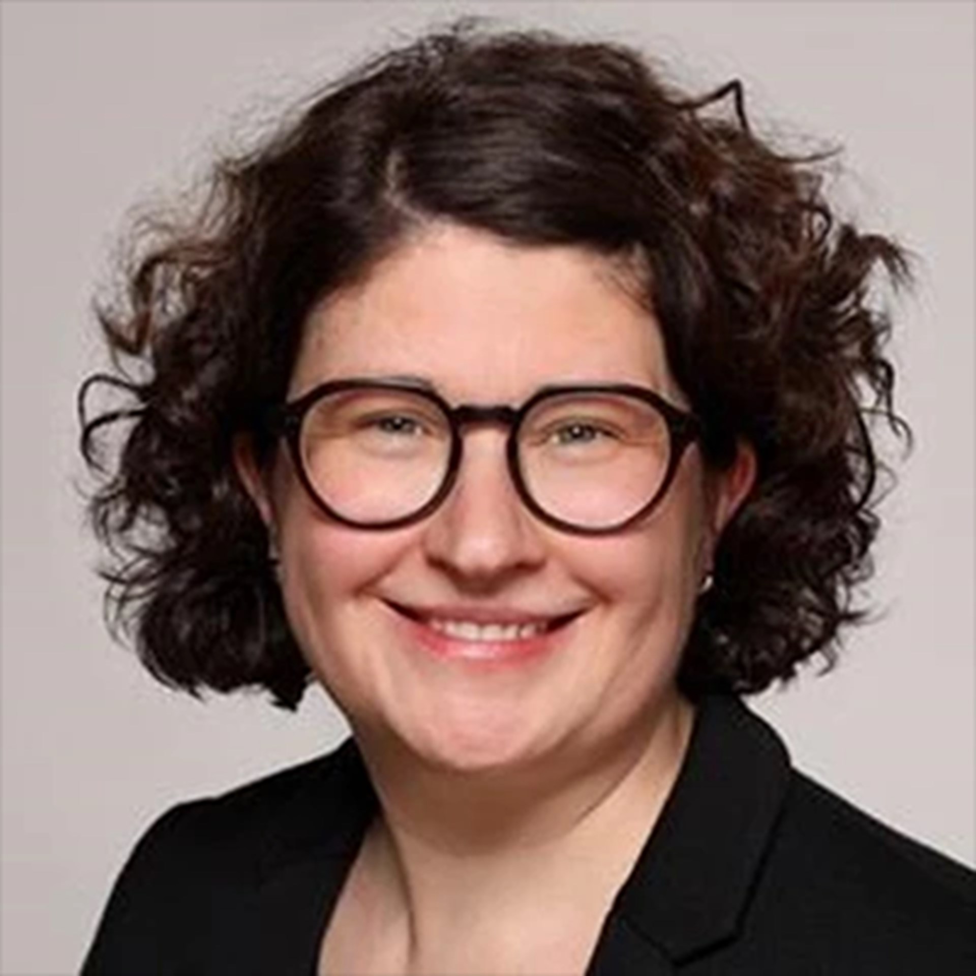 Lächelnde Frau mit braunen Haaren und Brille, die eine schwarze Anzugjacke trägt.
