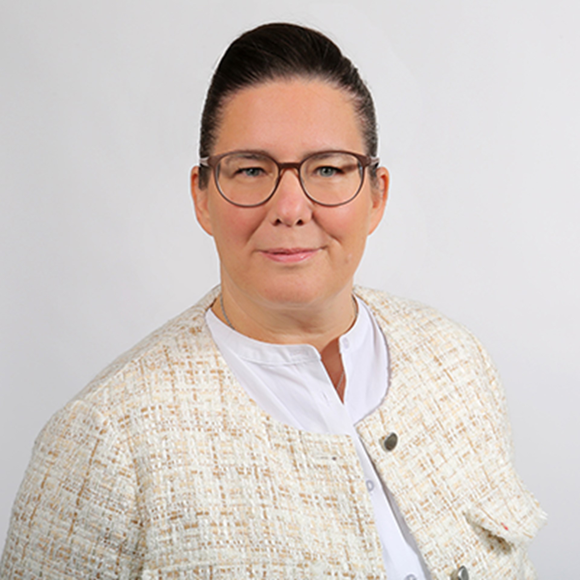 Sanft lächelnde Frau mit braunen Haaren und Brille, die ein weißes Shirt und eine helle, gemusterte Jacke trägt