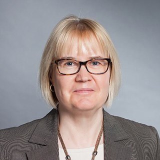 Frau mit blonden Haaren und Brille, die ein helles T-Shirt mit einer grauen Anzugjacke und einer Halskette trägt