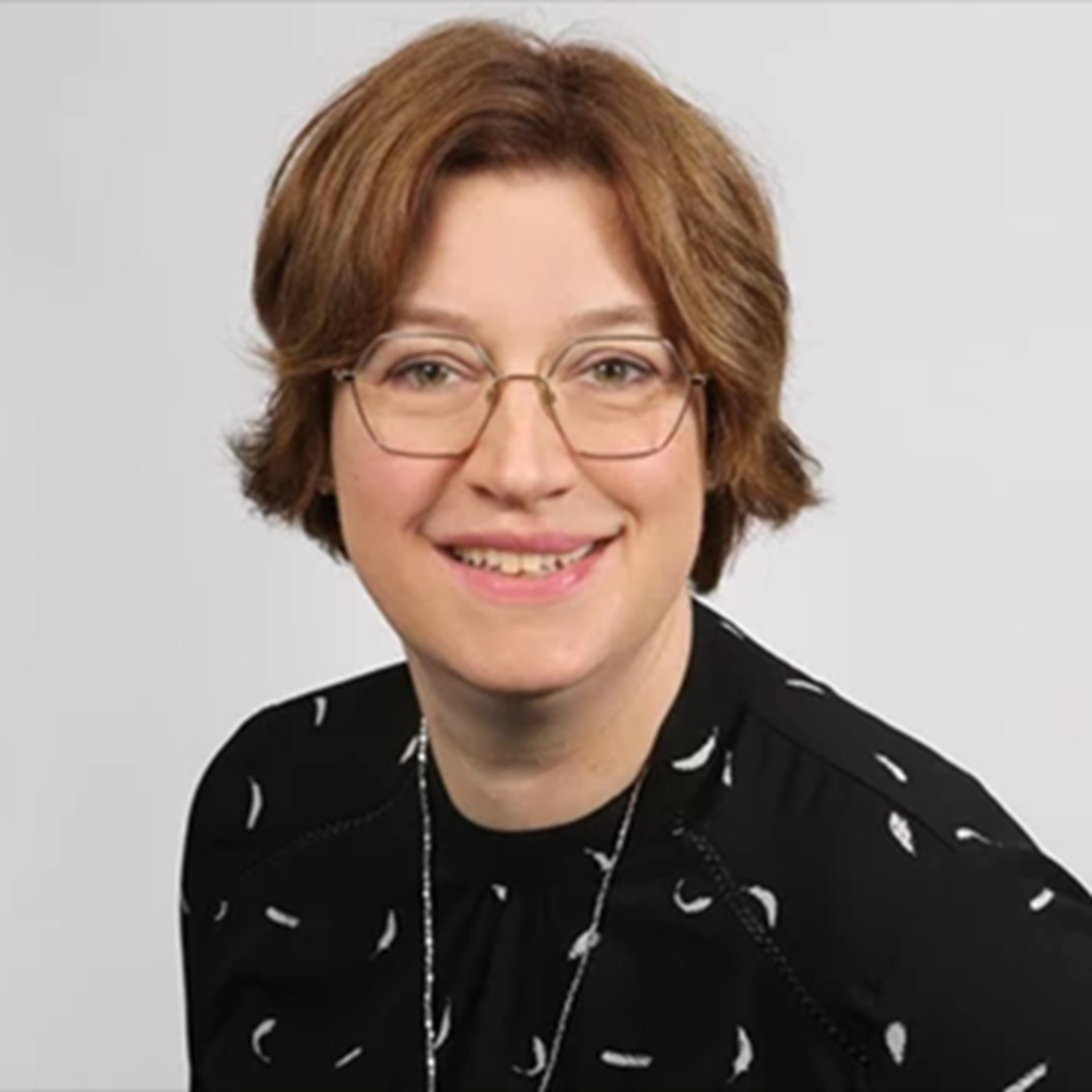 Lächelnde Frau mit braunen Haaren und Brille, die ein schwarzes Shirt mit weißem Muster und eine Kette trägt.