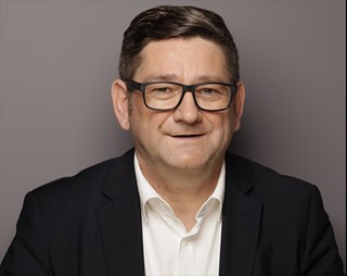 Lächelnder Mann mit braunen Haaren und Brille, der ein weißes Hemd und eine schwarze Anzugjacke trägt