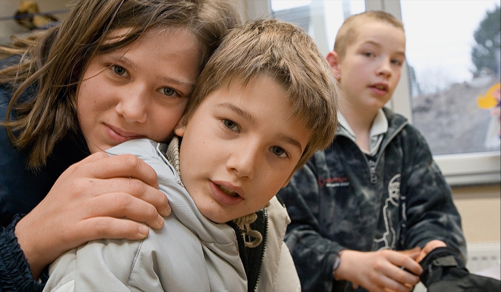 Ein Mädchen lehnt sich an den Rücken eines Jungen an, beide blicken in die Kamera. Im Hintergrund sitzt ein weiterer Junge.