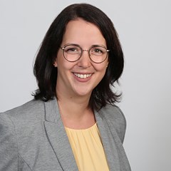 Lächelnde Frau mit schulterlangen, braunen Haaren und Brille, die ein gelbe Shirt und eine hellgraue Anzugjacke trägt