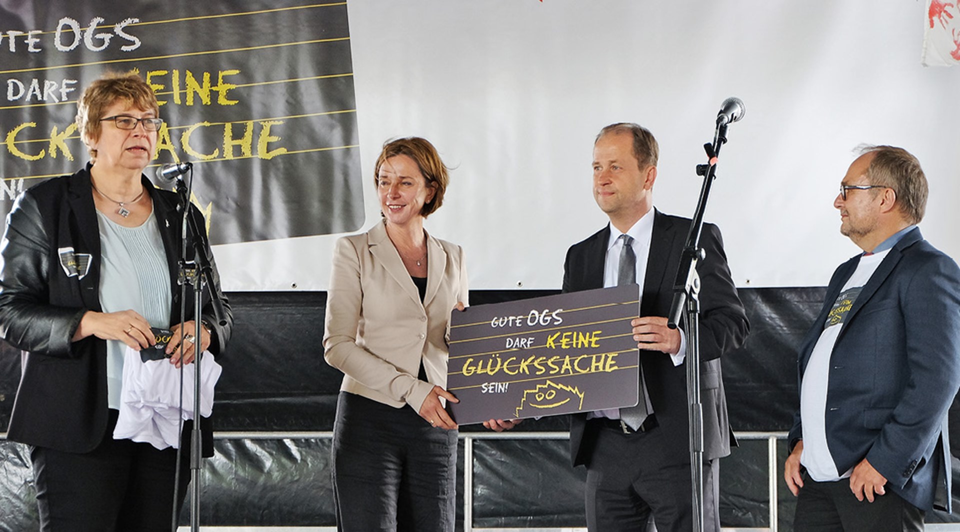 Zwei Frauen und zwei Männer stehen auf einer Bühne. Im Hintergrund und auf einem Schild ist das Logo der OGS-Kampagne 'Gute OGS darf keine Glücksache sein!' zu sehen.