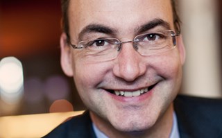 Lächelnder Mann mit braunen Haaren und Brille, der ein blaues Hemd mit einer dunklen Anzugjacke trägt und in einer Lokalität sitzt