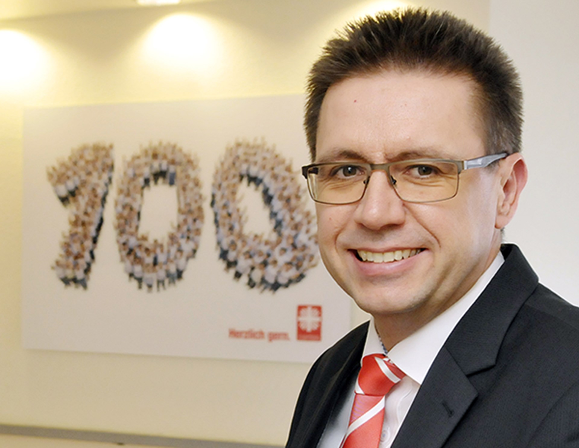 Lächelnder Mann mit blonden Haaren und Brille, der ein weißes Hemd, eine rot-weiß-gestreifte Krawatte und eine schwarze Anzugjacke trägt. Links neben ihm ist an einer Wand ein beleuchtetes Bild mit der Zahl 100 zu sehen.