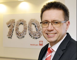 Lächelnder Mann mit blonden Haaren und Brille, der ein weißes Hemd, eine rot-weiß-gestreifte Krawatte und eine schwarze Anzugjacke trägt. Links neben ihm ist an einer Wand ein beleuchtetes Bild mit der Zahl 100 zu sehen.