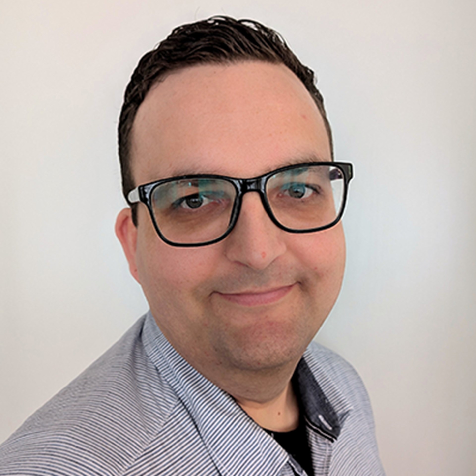 Lächelnder Mann mit schwarzen Haaren und Brille, der ein schwarzes T-Shirt und ein blau gestreiftes Hemd trägt.