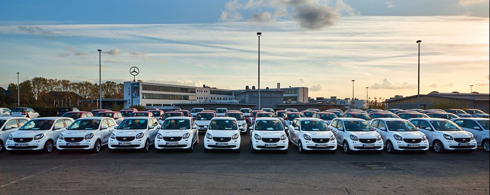Eine große Zahl an Elektro-Smarts, die aufgereiht auf einem Parkplatz stehen. Im Hintergrund ist ein Firmengebäude von Mercedes-Benz zu sehen.