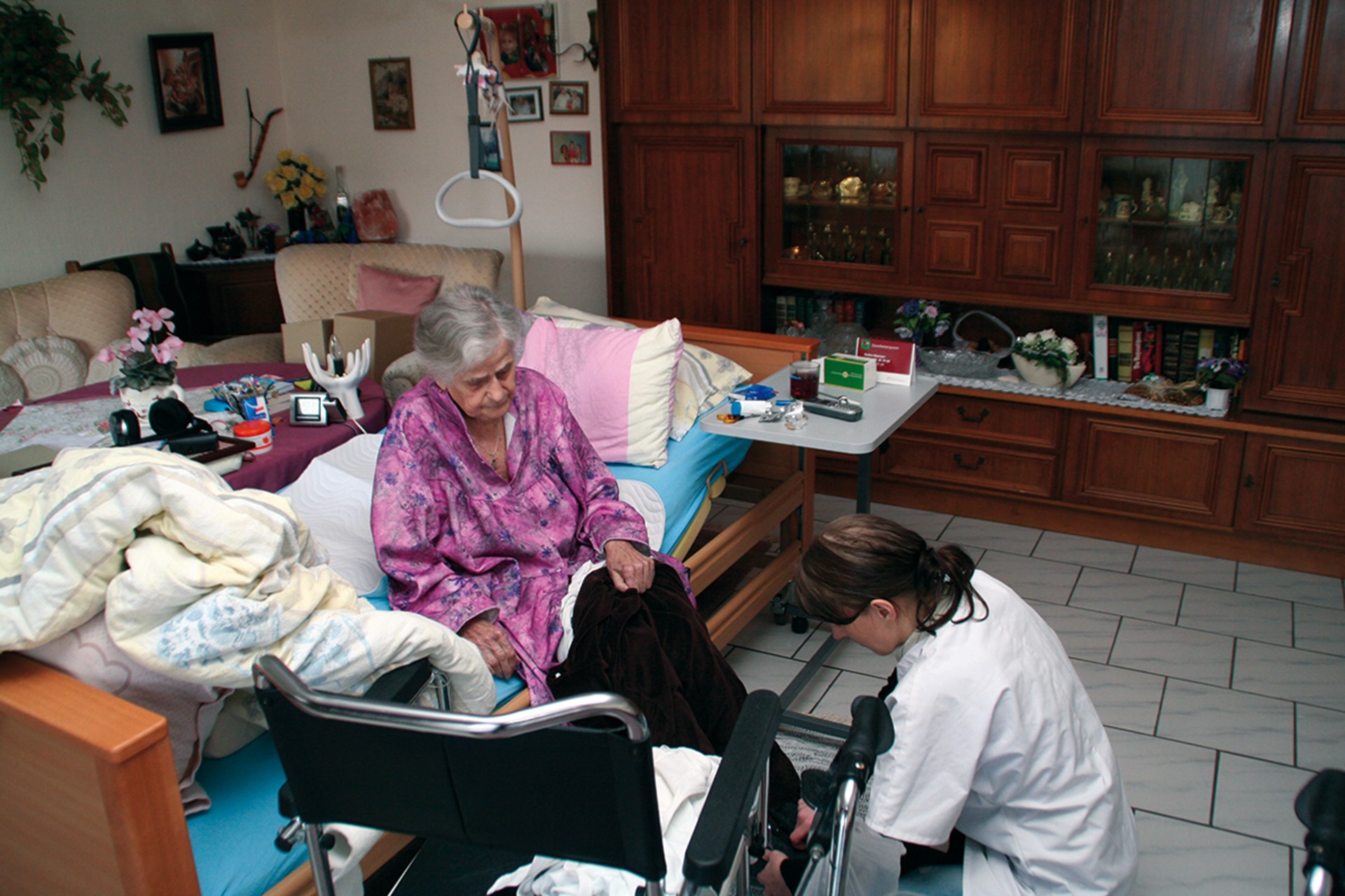Eine Seniorin sitzt auf einem Pflegebett, das in einem Wohnzimmer steht, und wird von einer Pflegerin, die vor ihr kniet, betreut