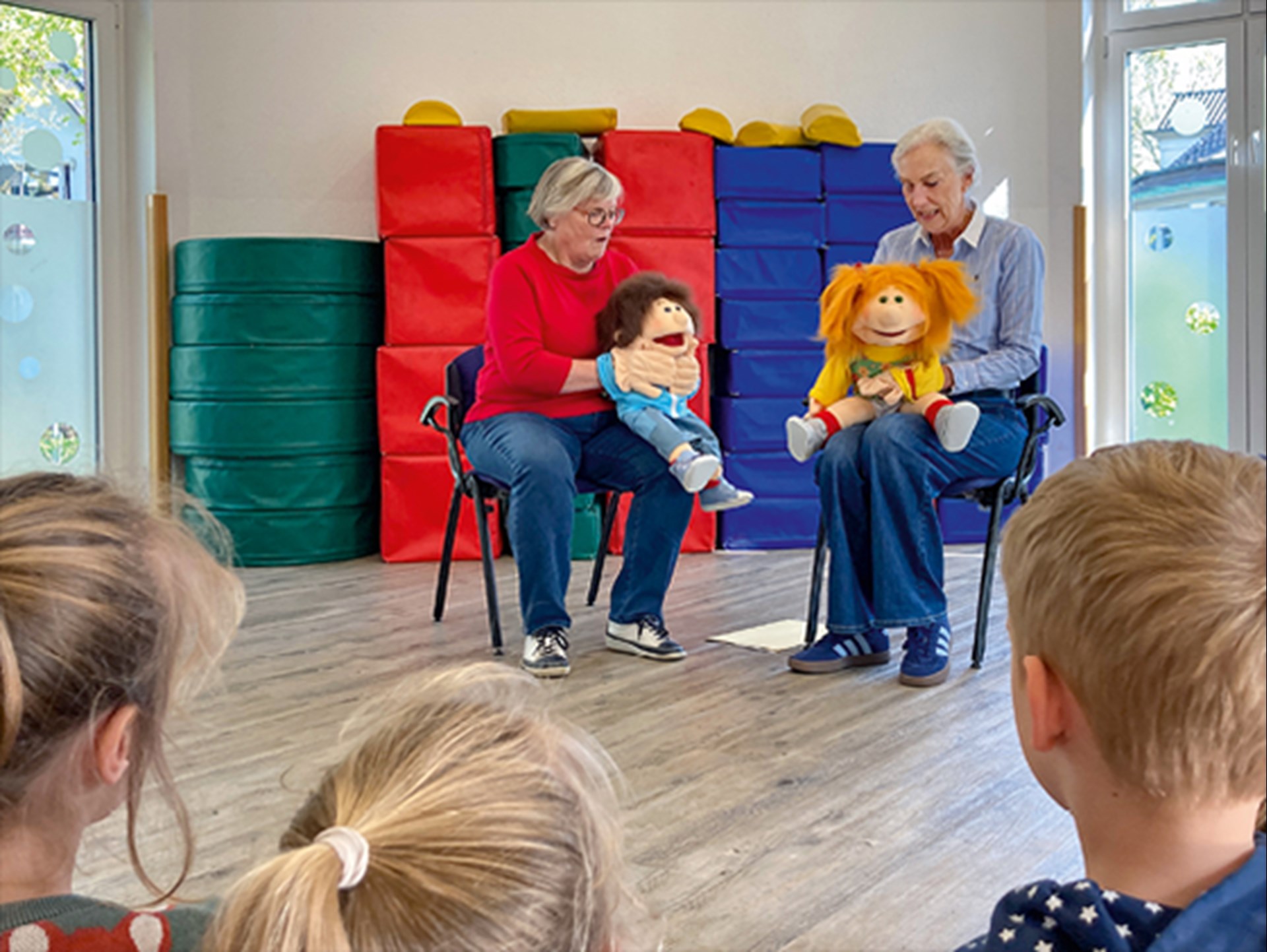 Zwei Ehrenamtliche der Malteser sitzen in einer Kindertagesstätte auf Stühlen und spielen mit großen Handpuppen eine Szene. Im Vordergrund sind die Hinterköpfe von zwei Mädchen und einem Jungen zu sehen.
