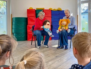 Zwei Ehrenamtliche der Malteser sitzen in einer Kindertagesstätte auf Stühlen und spielen mit großen Handpuppen eine Szene. Im Vordergrund sind die Hinterköpfe von zwei Mädchen und einem Jungen zu sehen.