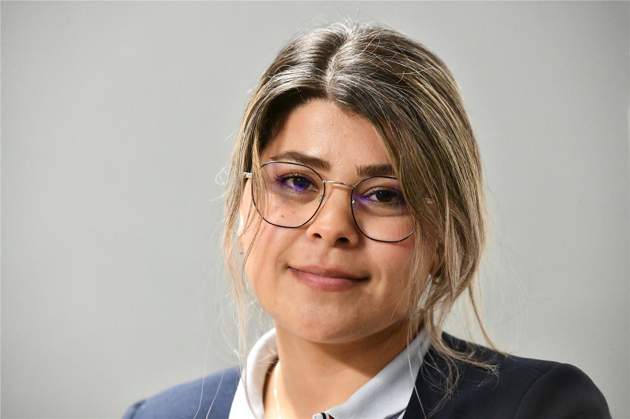 Lächelnde Frau mit blonden Haaren und Brille, die ein weißes Hemd und eine blaue Anzugjacke trägt