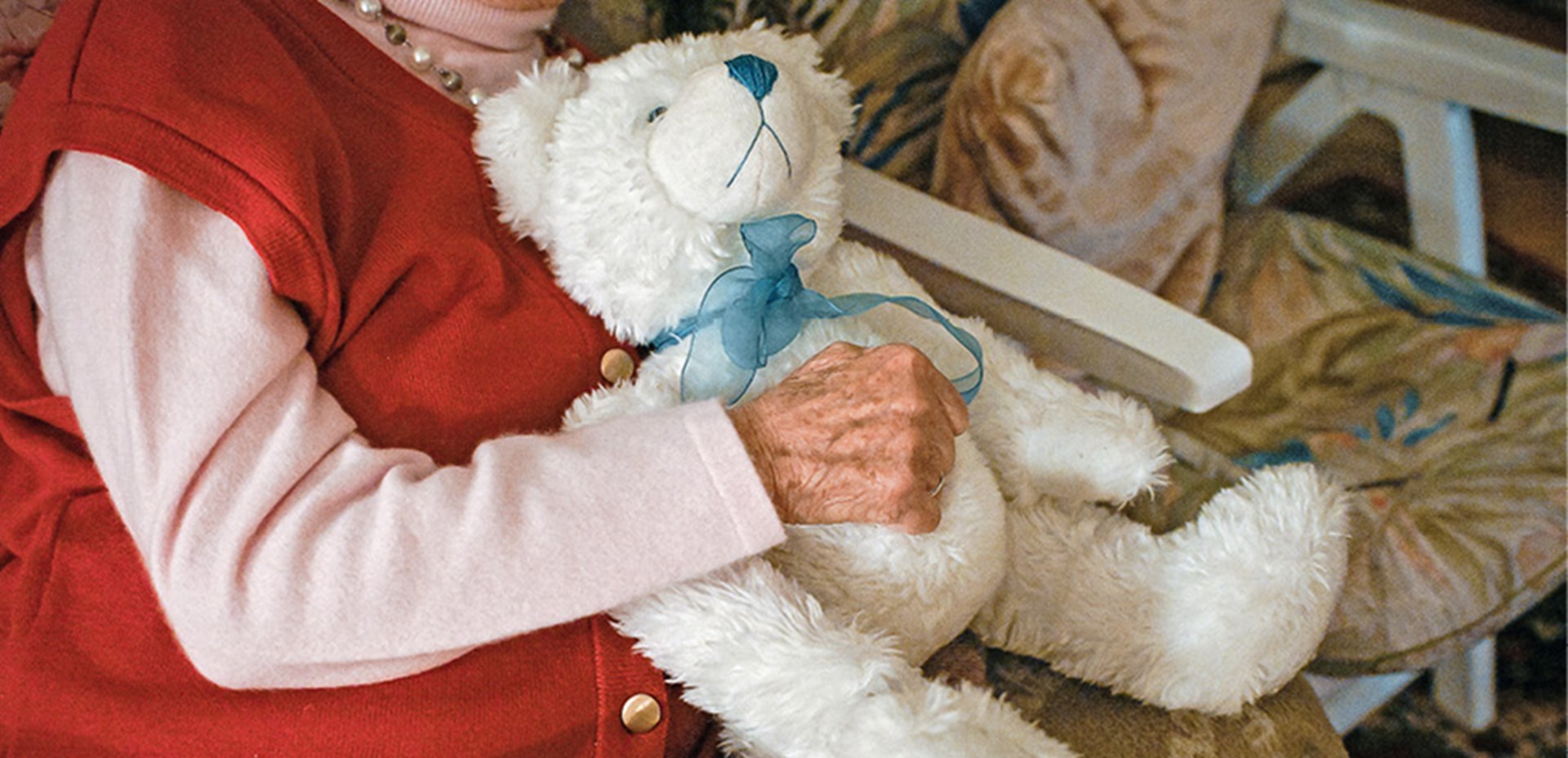 Eine Seniorin, die auf einem Stuhl sitzt und einen weißen Teddybär mit blauer Schleife im Arm hält