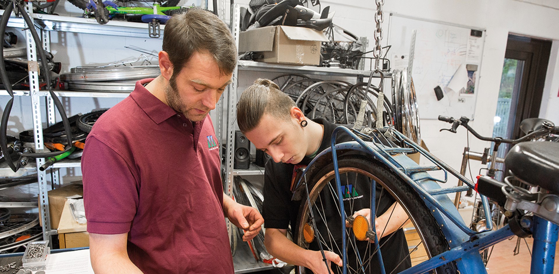Zwei Männer stehen in einer Werkstatt und arbeiten an einem Fahrrad. Im Hintergrund sind einige Metallregale mit Ersatzteilen zu sehen.