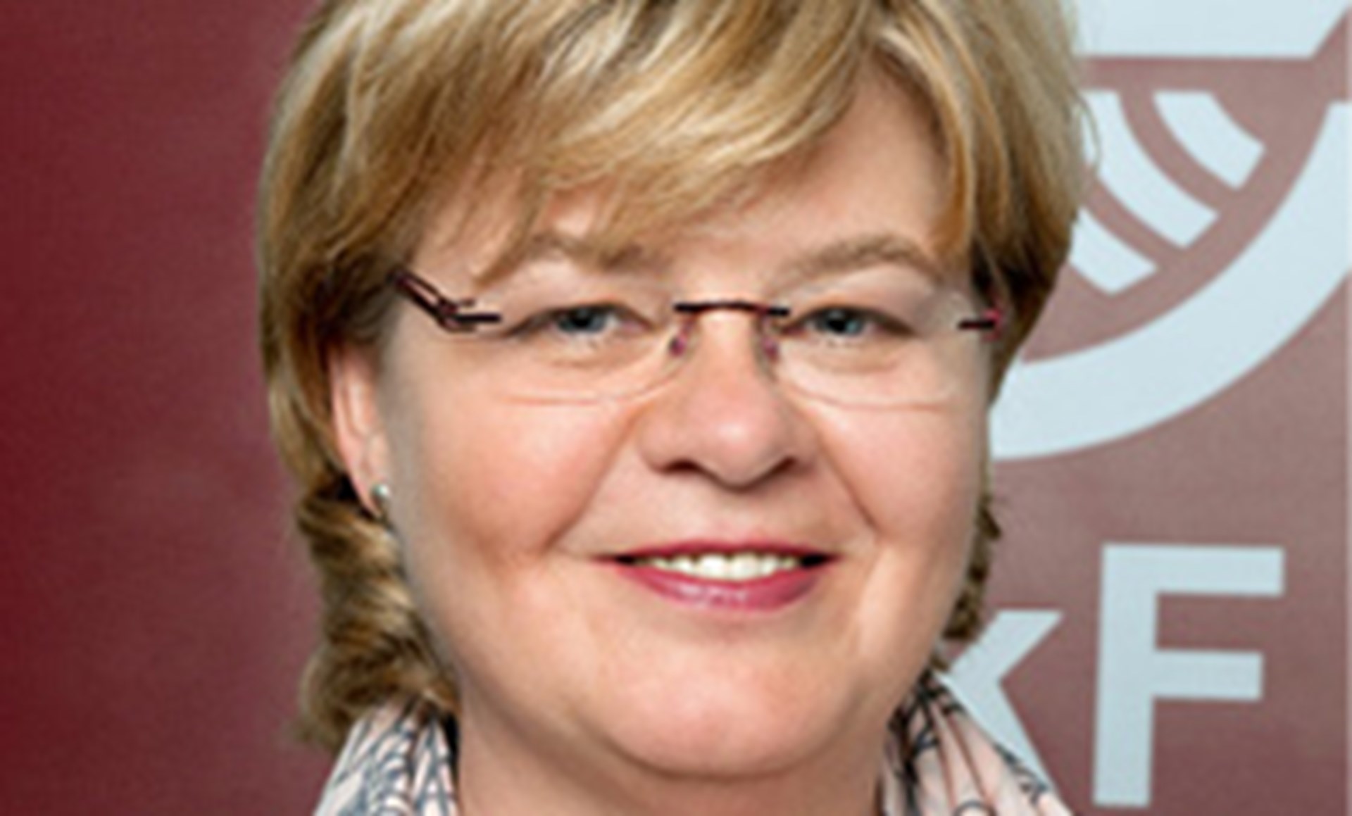 Lächelnde Frau mit blonden Haaren und Brille, die einen hellen Schal trägt und vor einem roten Hintergrund mit einem weißen SkF-Logo steht