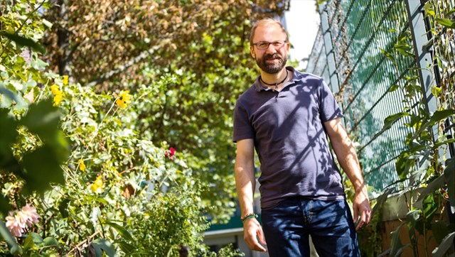 Ein vollbärtiger Mann, der ein lila Polohemd mit einer blauen Jeans trägt und auf einem dicht bewachsenen Gelände neben einem grünen Zaun steht. Er wird von der Sonne angestrahlt.