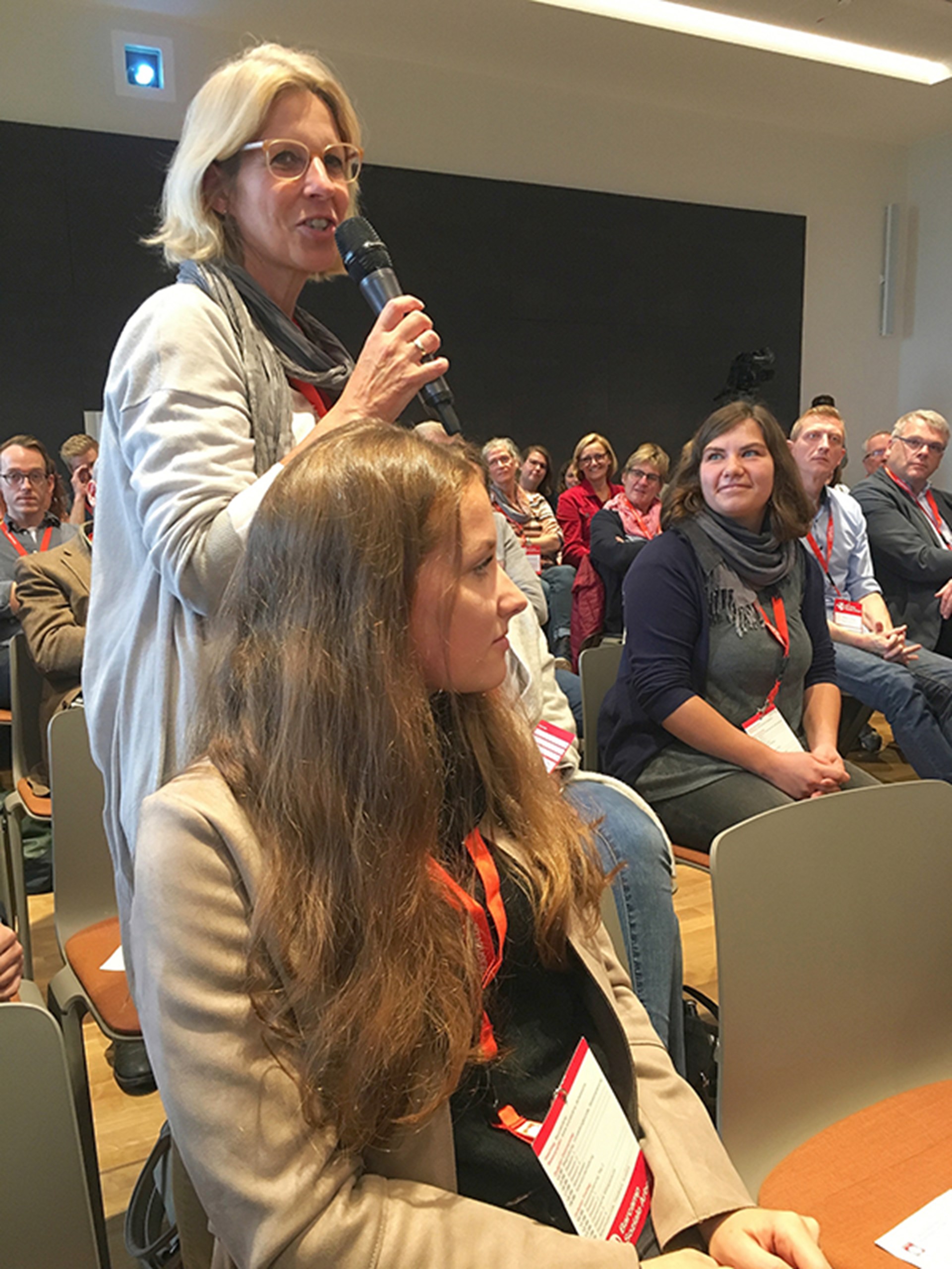 Eine Frau aus dem Publikum des Barcamps Soziale Arbeit in Siegburg steht vor ihrem Stuhl und hält ein Mikrofon in der Hand