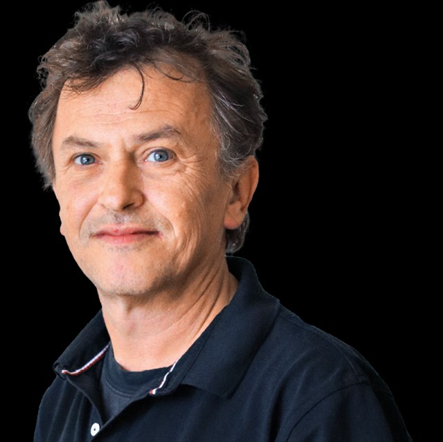 Lächelnder Mann mit grau-braunen Haaren, der ein blaues T-Shirt und einen blauen Pullover trägt