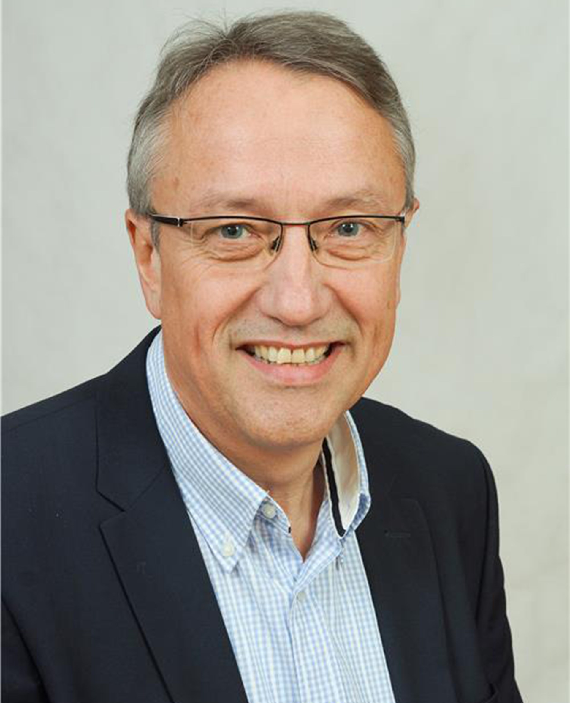 Lächelnder Mann mit grauen Haaren und Brille, der ein blaues Hemd und eine dunkelblaue Anzugjacke trägt