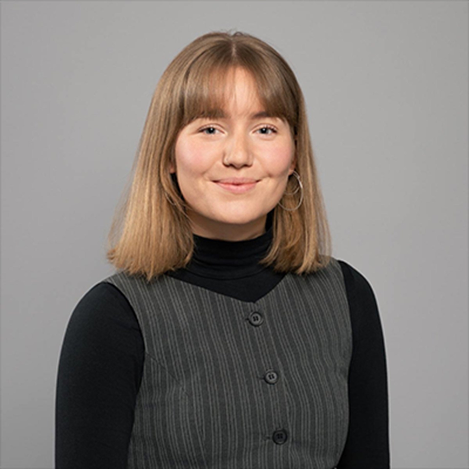 Lächelnde Frau mit schulterlangen, blonden Haaren, die einen schwarzen Pullover und eine graue Weste trägt.