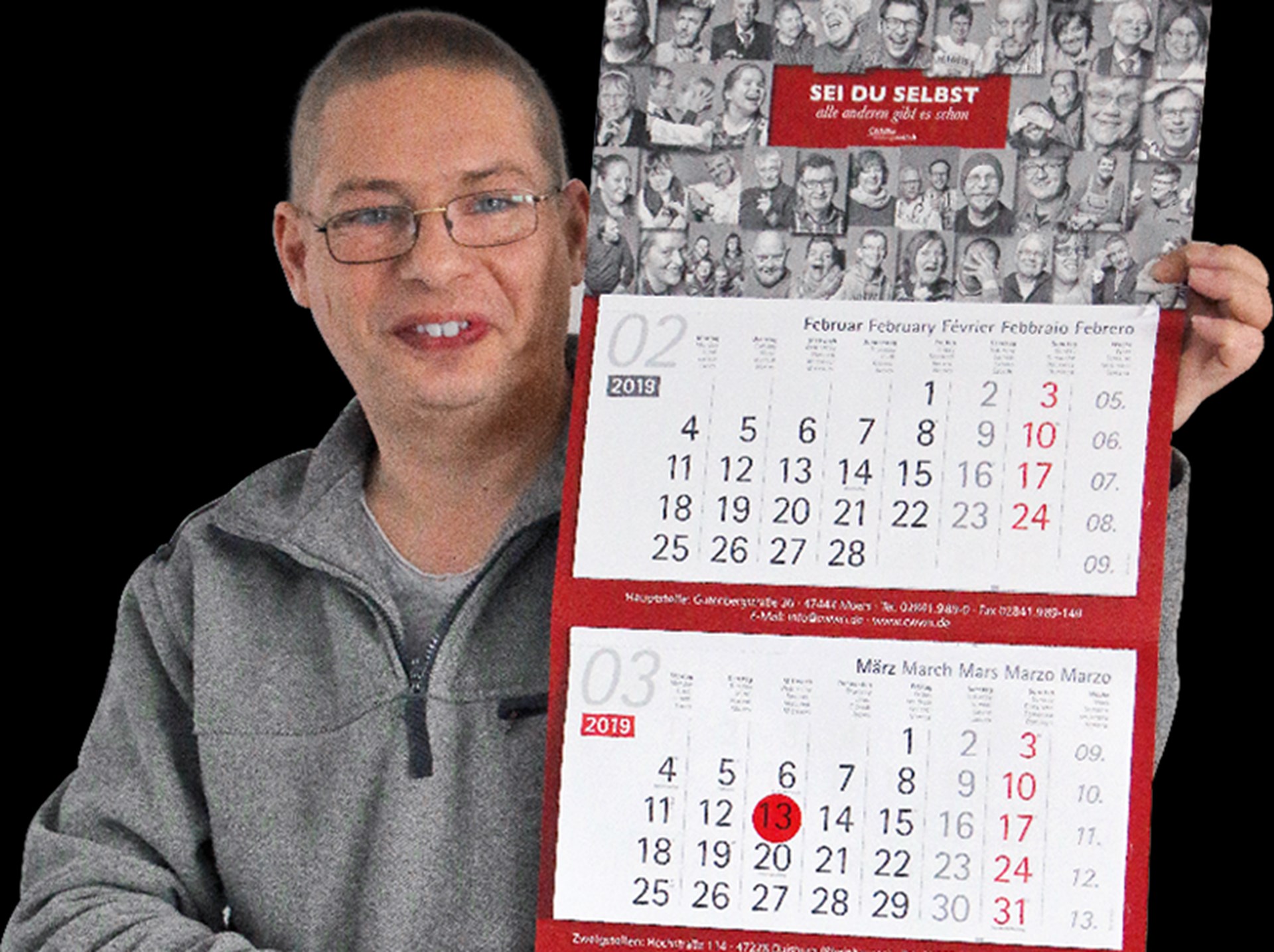 Lächelnder Mann mit kurzen Haaren und Brille, der eine graue Jacke trägt und einen selbst produzierten Dreimonatskalender hoch hält
