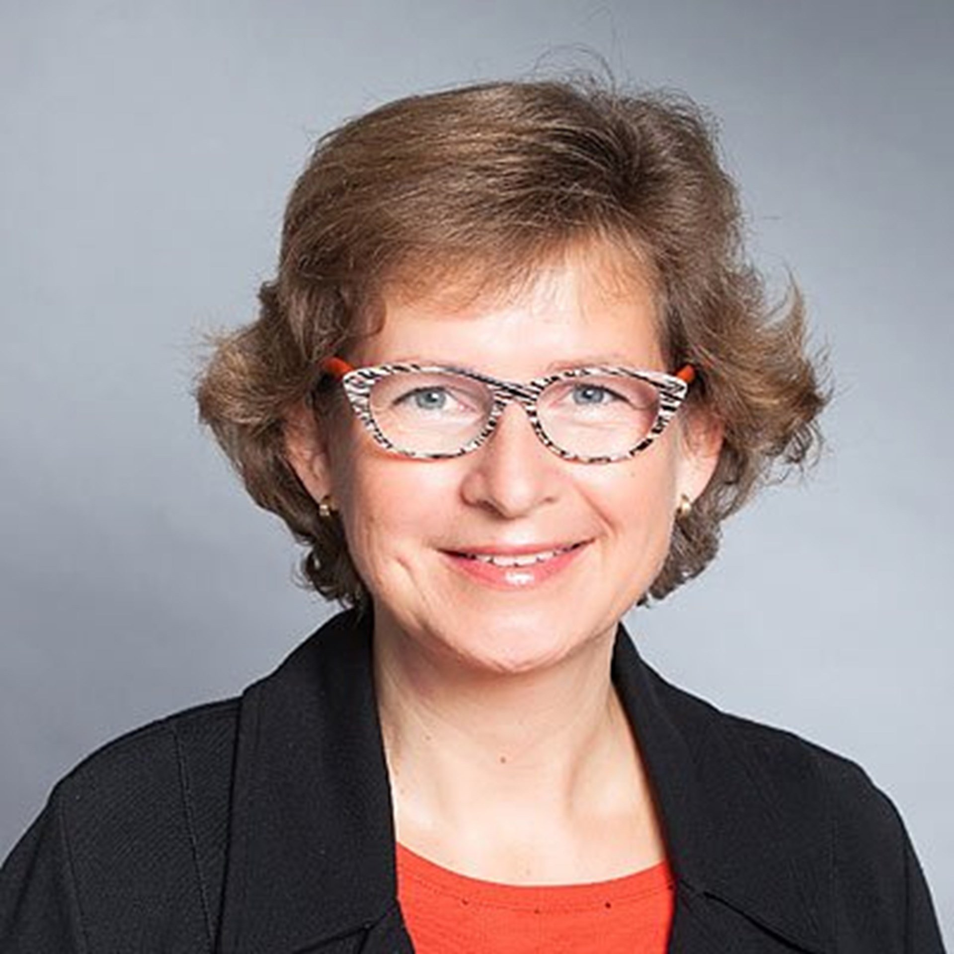 Lächelnde Frau mit blonden Haaren und einer bunten Brille, die ein rotes T-Shirt und eine schwarze Anzugjacke trägt
