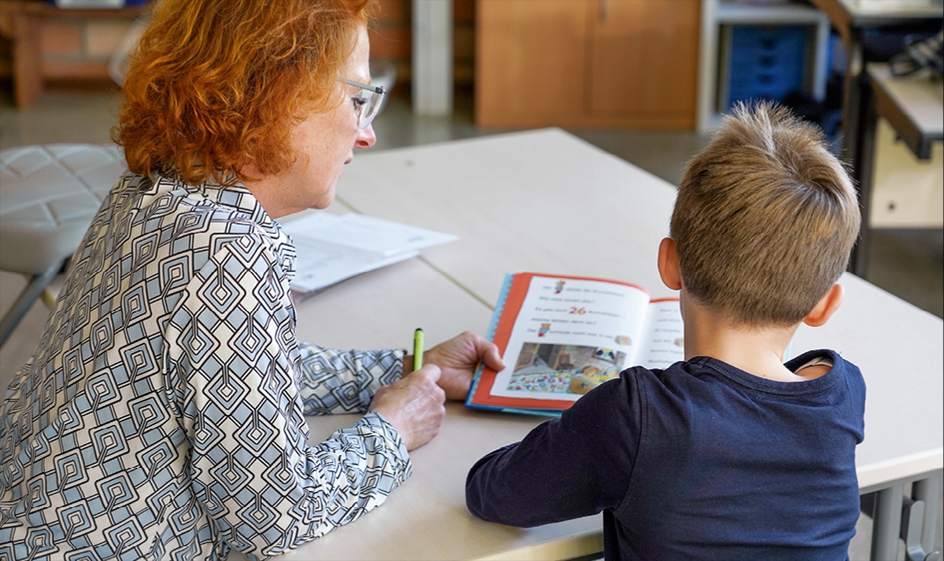 Eine Frau mit roten Haaren sitzt neben einem Jungen an einem Tisch und liest mit ihm ein Buch. Beide sitzen mit dem Rücken zur Kamera.