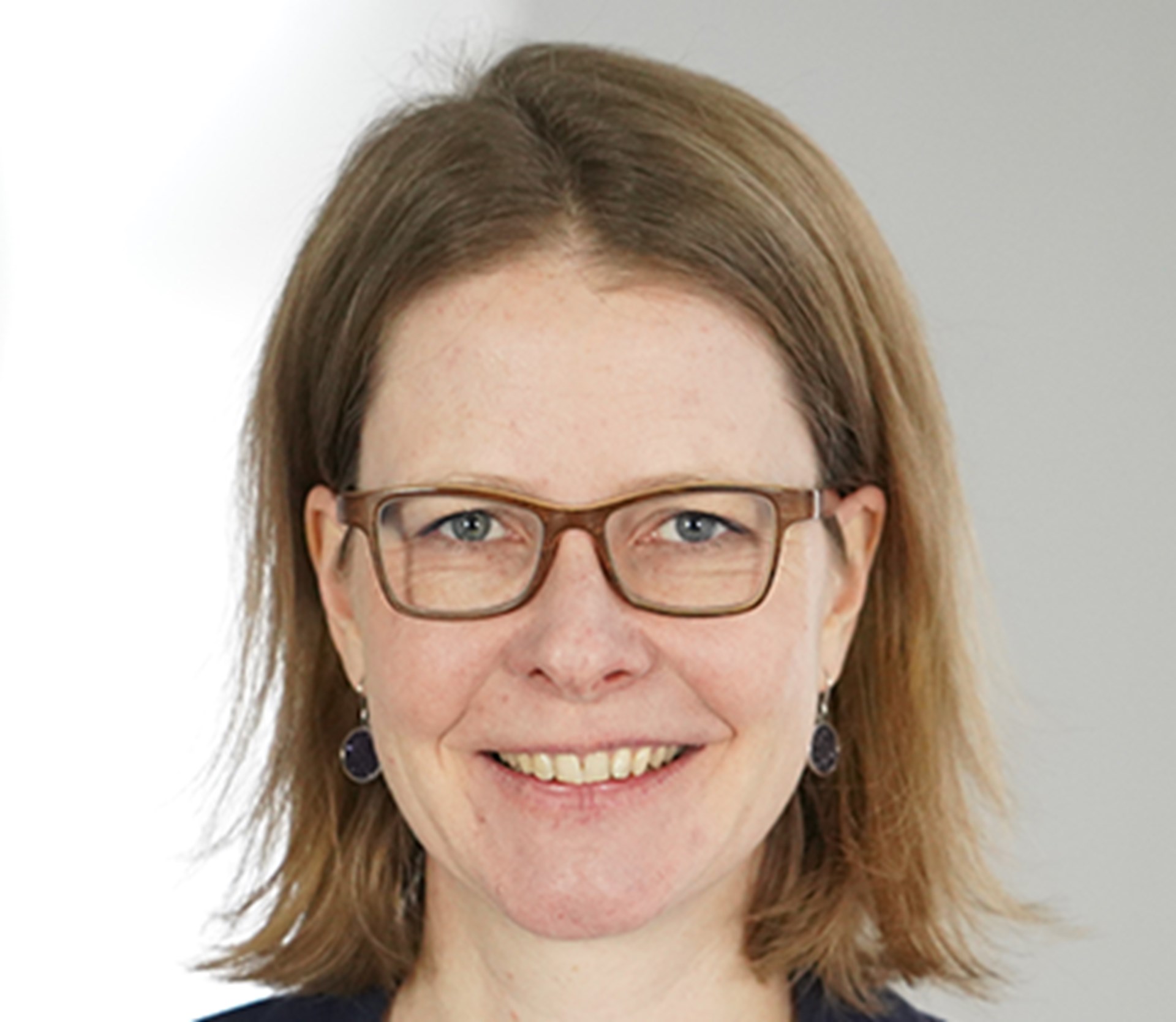 Lächelnde Frau mit langen, braunen Haaren und blauer Brille, die ein weißes Shirt und eine blaue Anzugjacke trägt