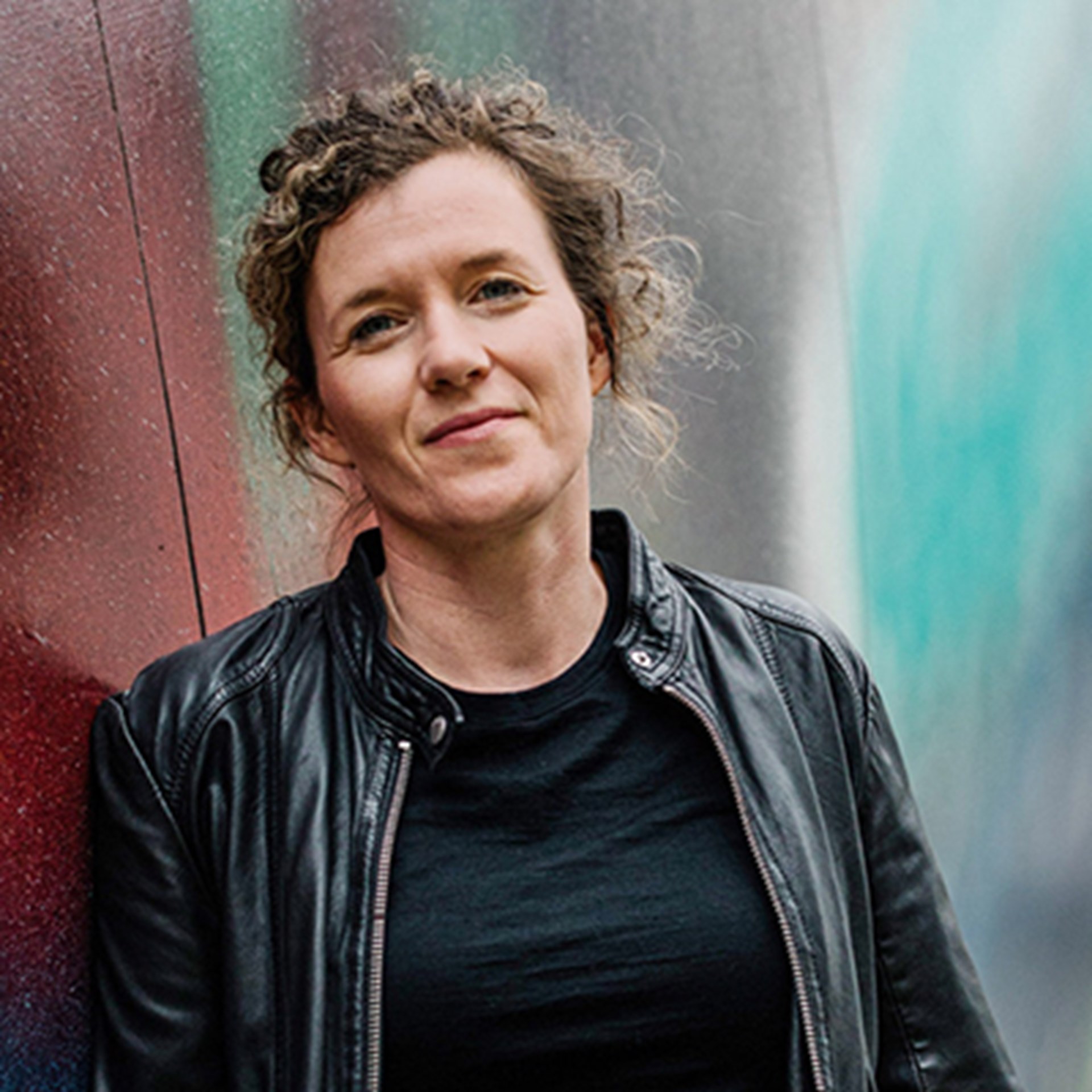 Frau mit lockigem, brauen Haaren, die ein schwarzes T-Shirt mit einer schwarzen Lederjacke trägt und an eine mit Graffiti besprühten Wand lehnt