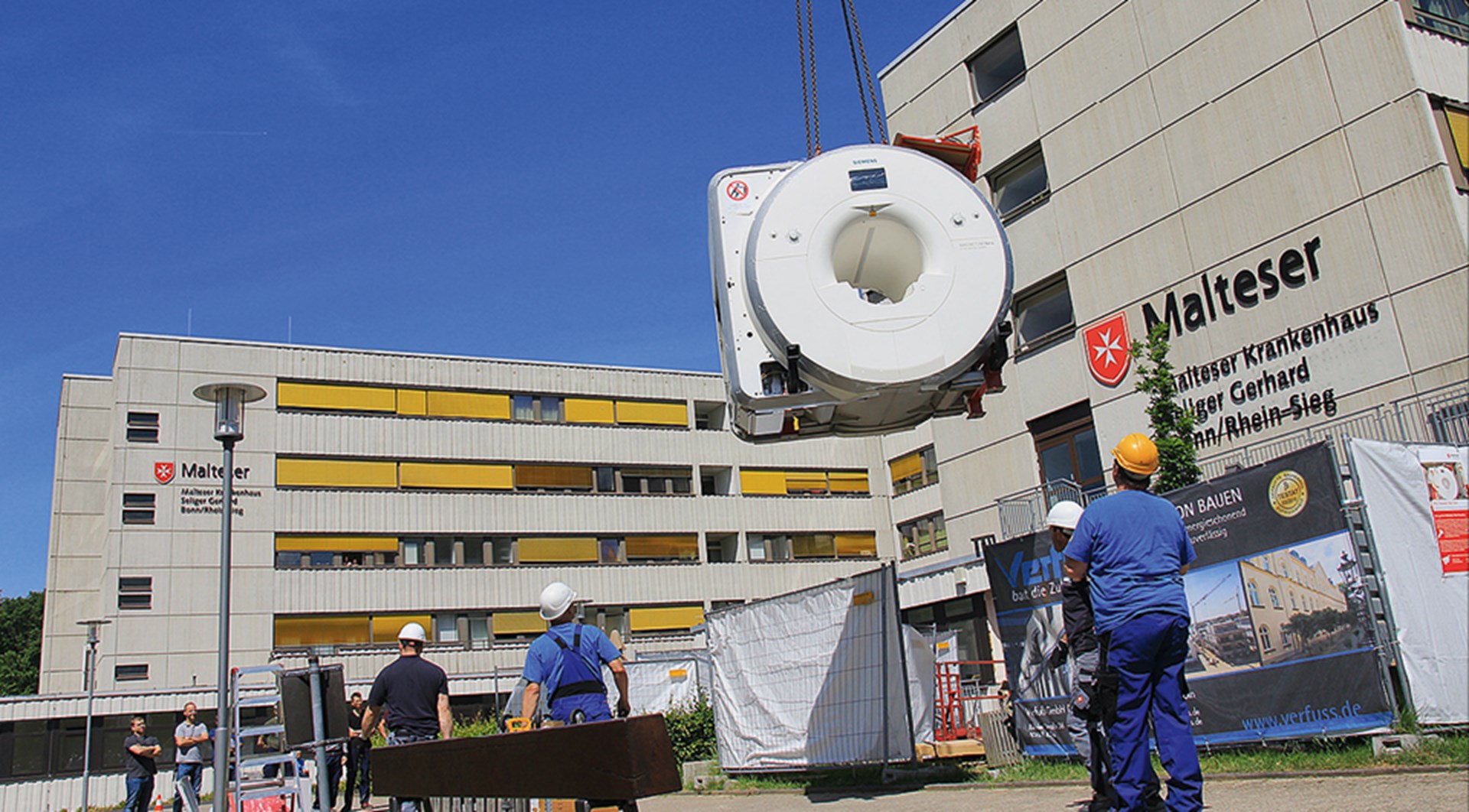 Einige Bauarbeiter stehen an einer Baustelle vor dem Malteser Krankenhaus 'Seliger Gerhard' Bonn/Rhein-Sieg und beobachten, wie ein Kran ein großes Objekt transportiert