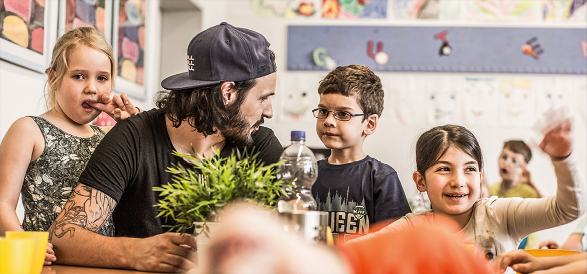 Ein Erzieher sitzt in einer Offenen Ganztagsschule an einem Tisch. Um ihn herum stehen und sitzen einige Kinder.
