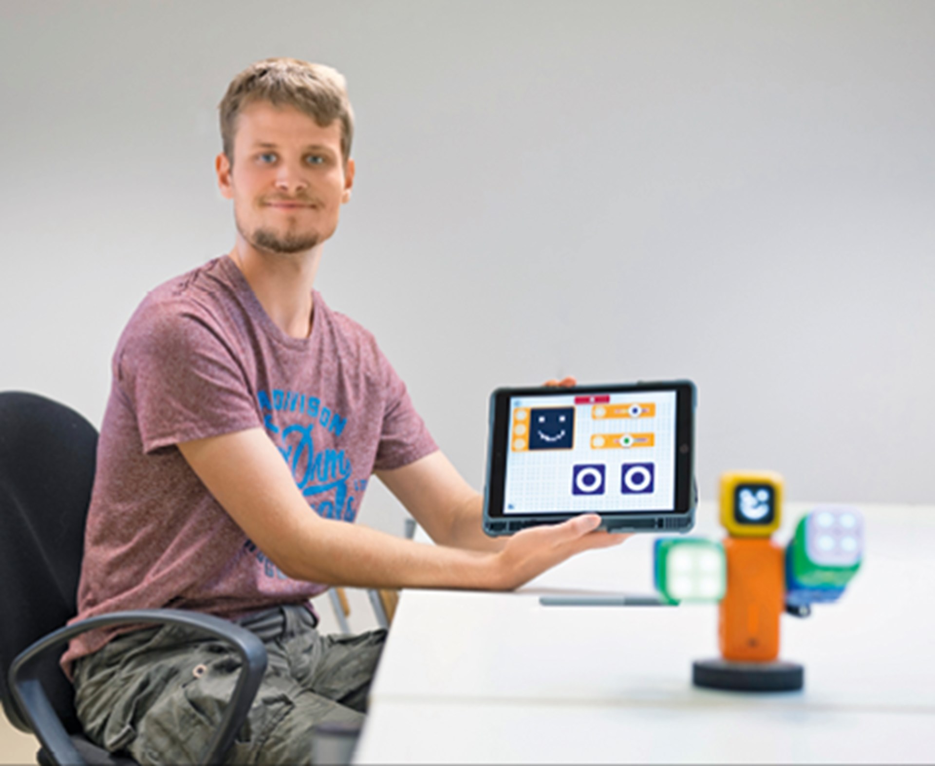 Ein Mann sitzt in einem Bürostuhl an einem Tisch und hält ein Tablet in die Kamera, auf dem einem Anwendung ausgeführt wird