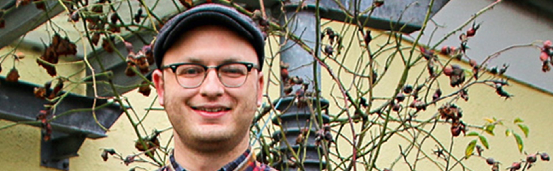 Lächelnder Mann mit Schiebermütze und Brille, der ein kariertes Hemd trägt und vor dem Eingang der St. Ida in Bockum-Hövel steht