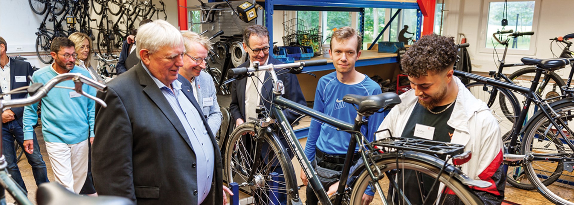 Ein Minister steht mit zwei Caritas-Vertretern in einer Fahrrad-Werkstatt des Schloss Bellinghoven und schaut Auszubildenden bei der Arbeit zu