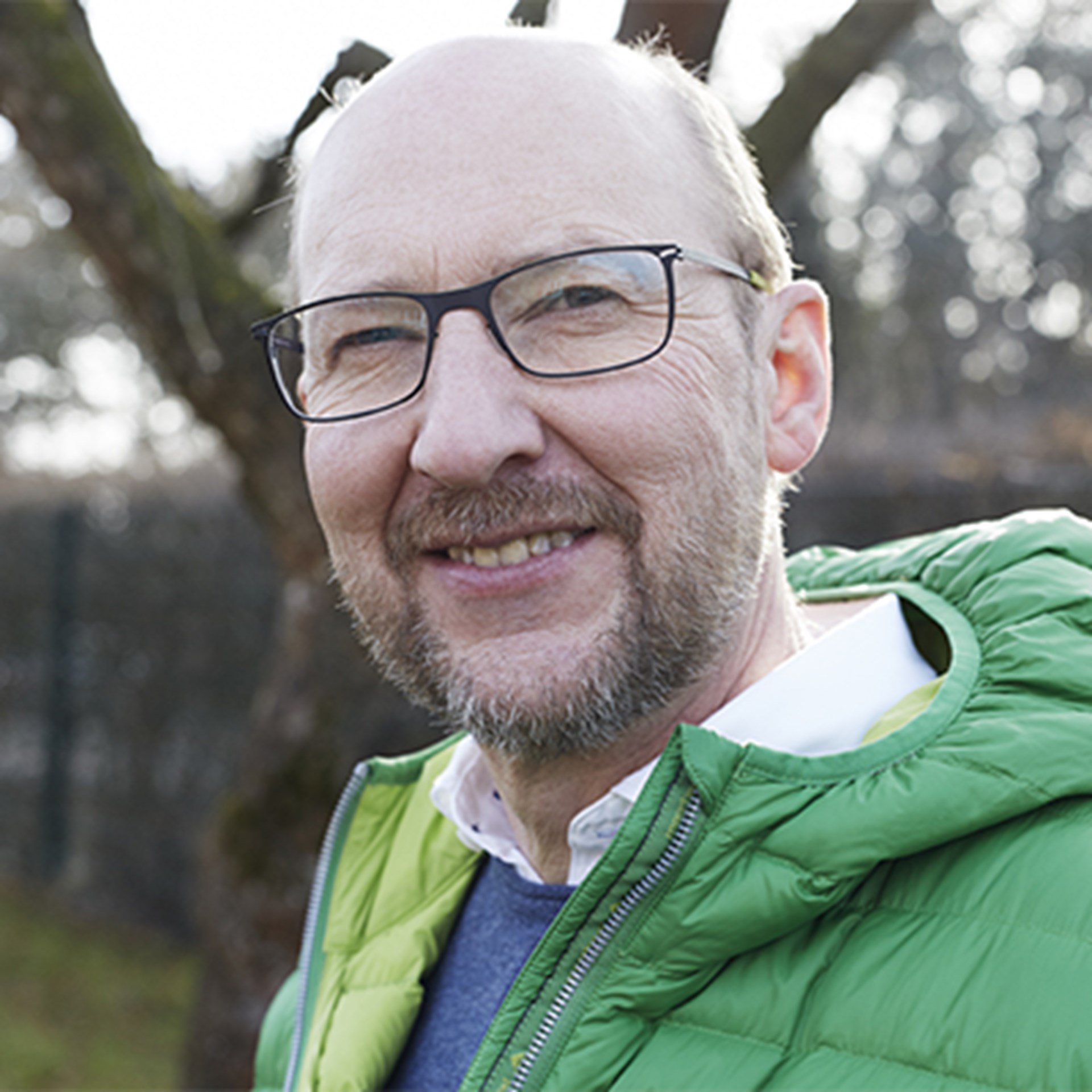 Lächelnder Mann mit Glatze und Vollbart, der eine grüne Jacke trägt und sich in einer Grünanlage befindet