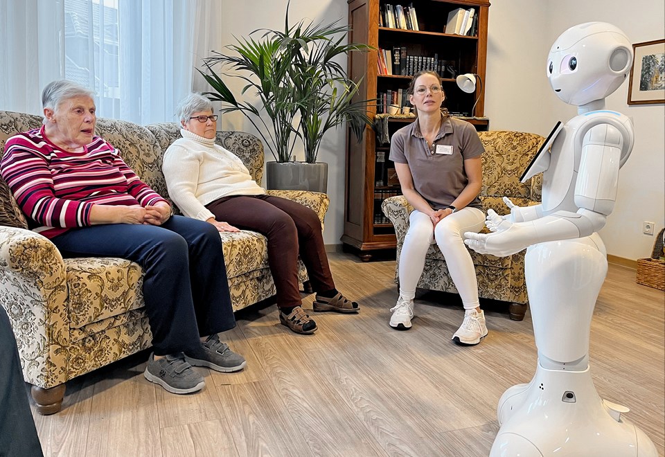 Zwei Seniorinnen und eine Pflegerin sitzen auf Polstermöbeln in einem Gemeinschaftsraum zusammen und blicken auf den Roboter Pepper
